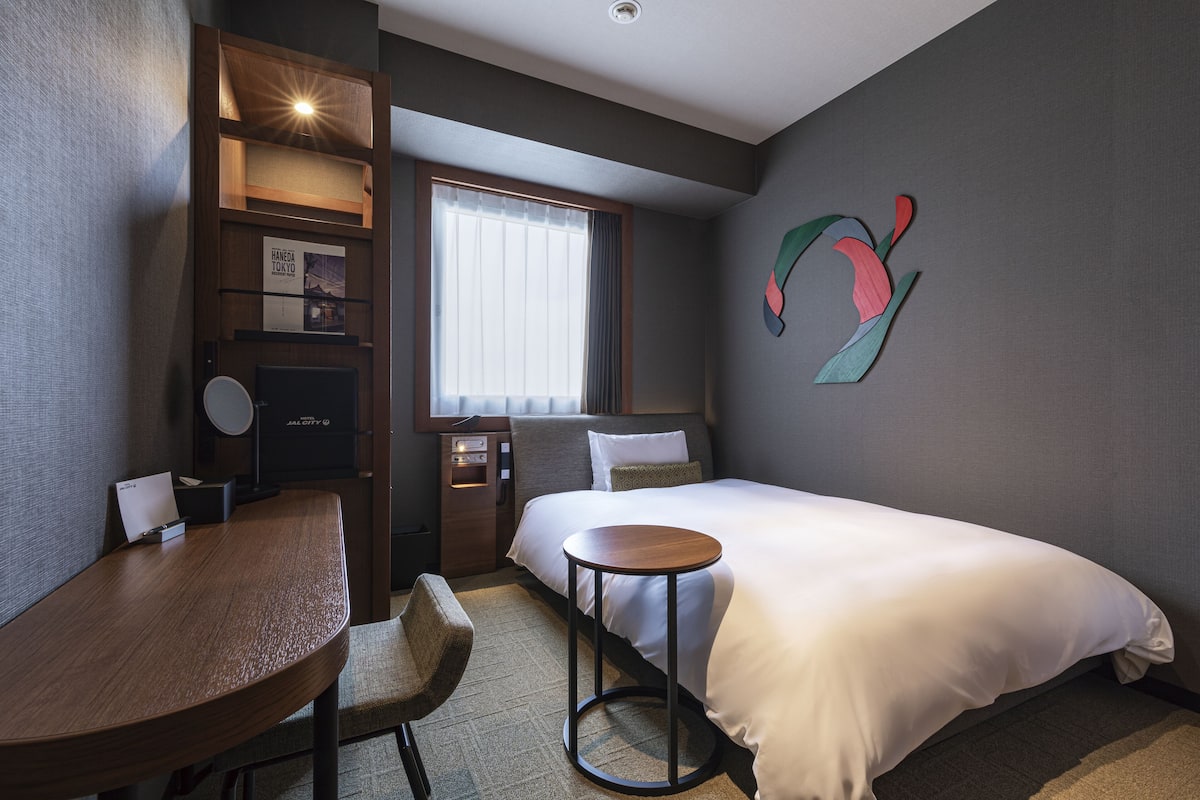A hotel room with a bed, a round table, a chair, a desk, a window with curtains, a wall-mounted artwork, and a shelf with a lamp and a framed picture.