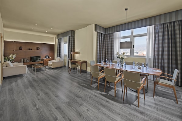 Una habitación de hotel moderna con comedor, un conjunto de sofás, un televisor y un amplio ventanal con cortinas.