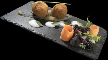 A slate serving board with two round, breaded items, a small dollop of cream, and a decorative arrangement of berries and greens.