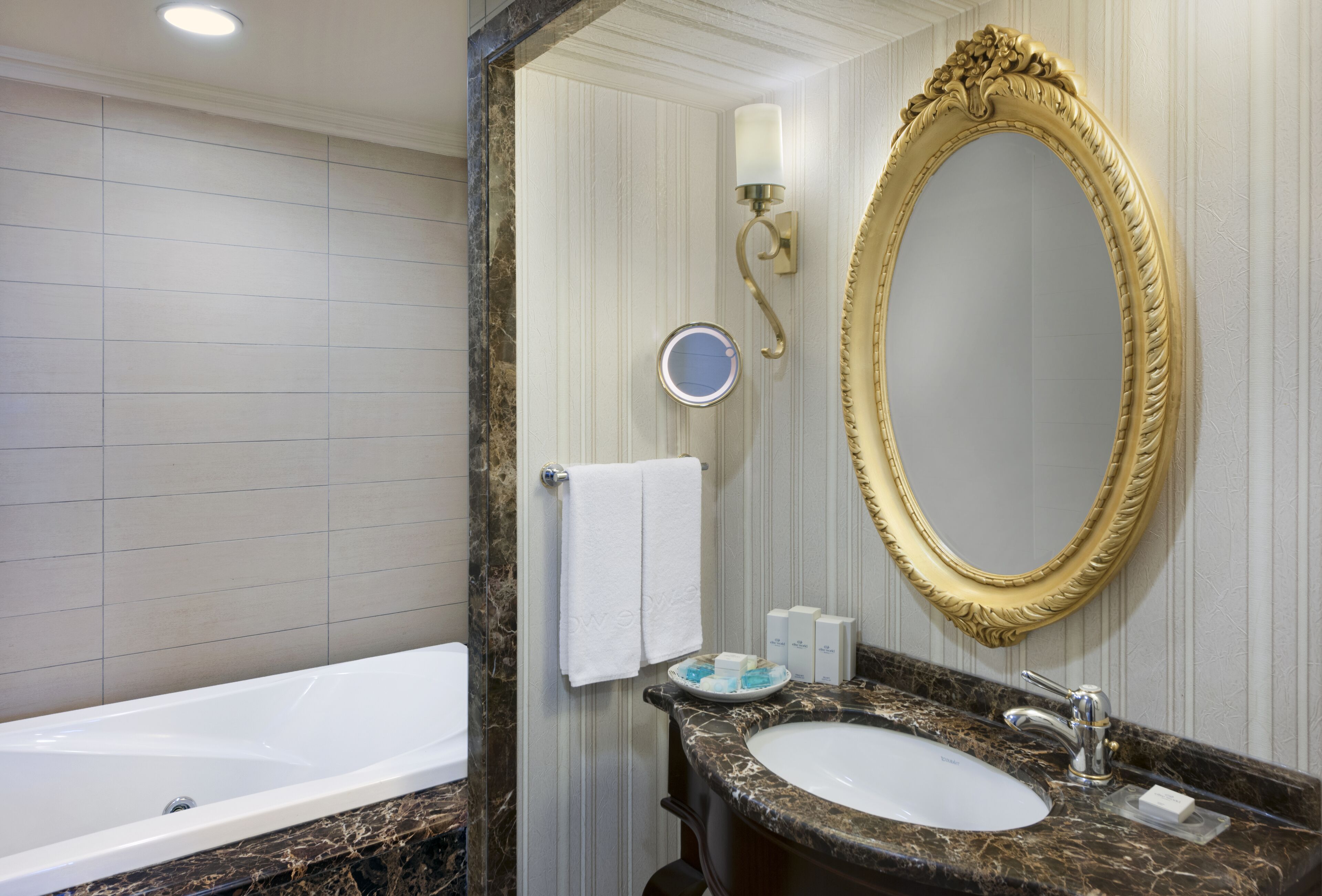 A bathroom with a bathtub, a sink with a marble countertop, a gold-framed mirror, and a wall-mounted light fixture.