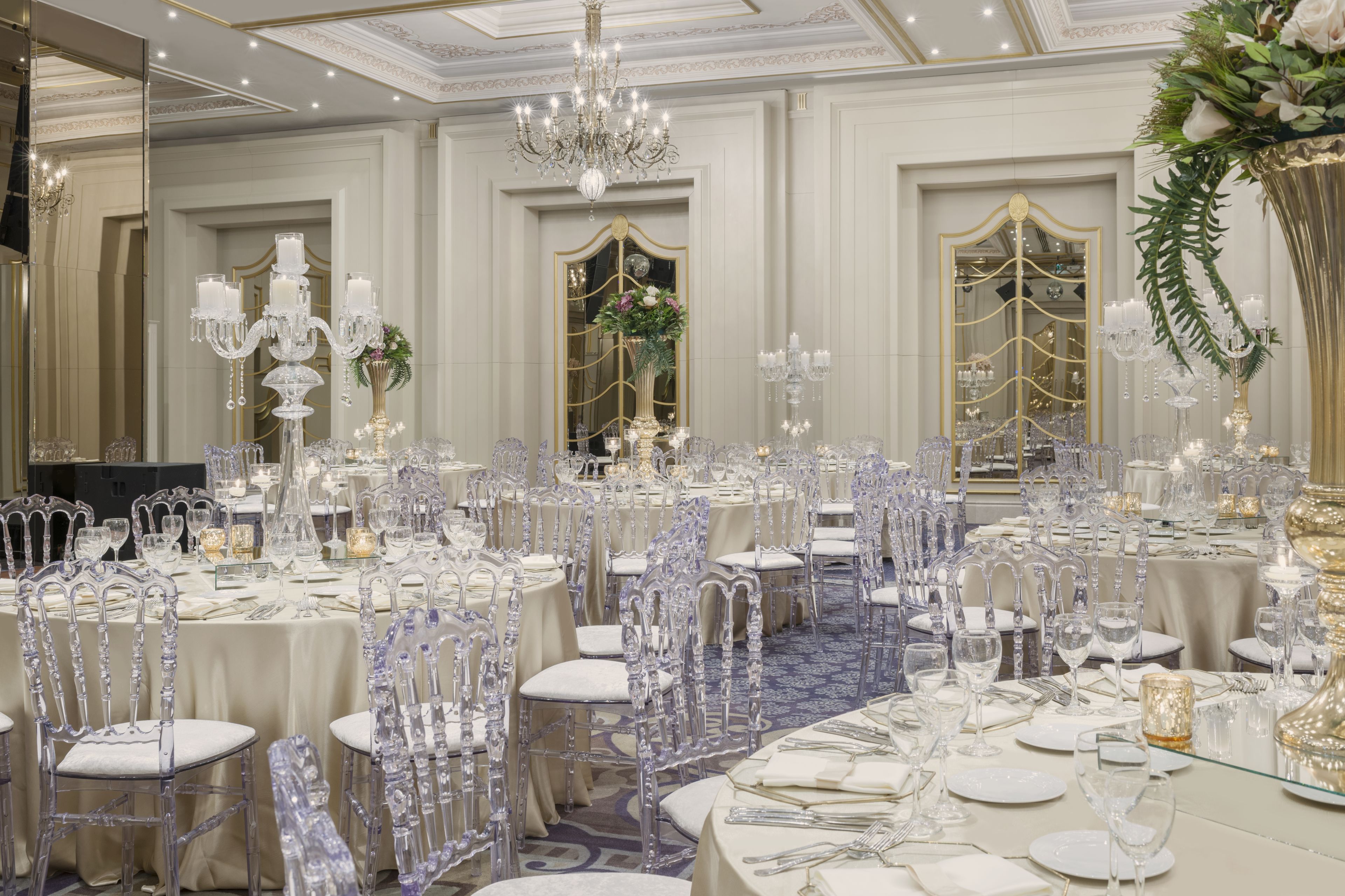 A banquet hall with round tables, clear chairs, and chandeliers.