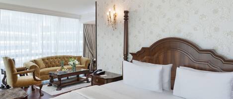 A hotel room with a large bed, a sofa, a wooden nightstand, a lamp, and a window with sheer curtains.