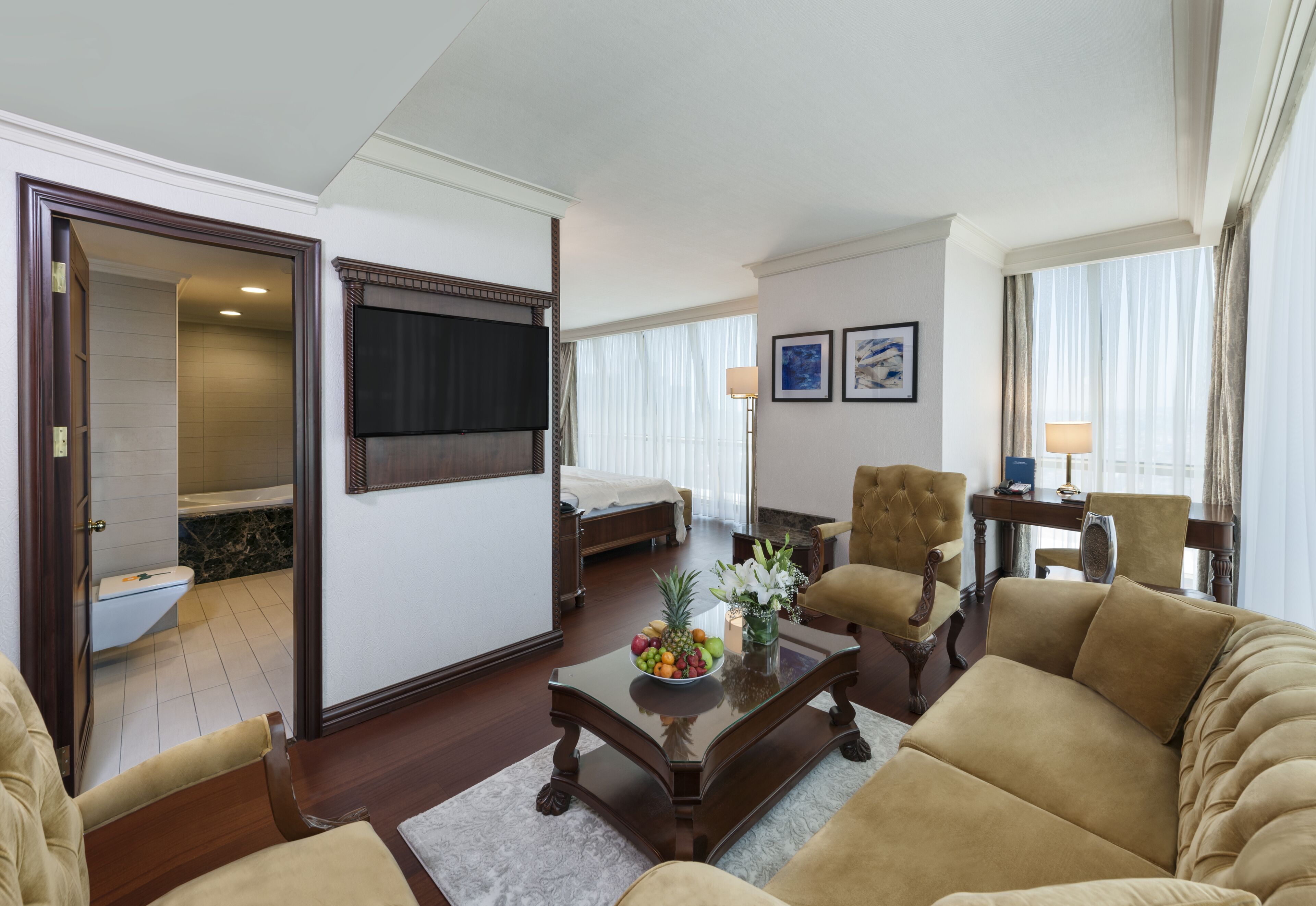 A hotel room with a living area featuring a sofa, armchairs, a coffee table with fruit, a flat-screen TV, and a bed with a white headboard.