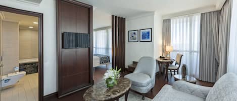 A hotel room with a sofa, armchair, and a small table with a vase of flowers. There is a television mounted on the wall and a window with curtains.