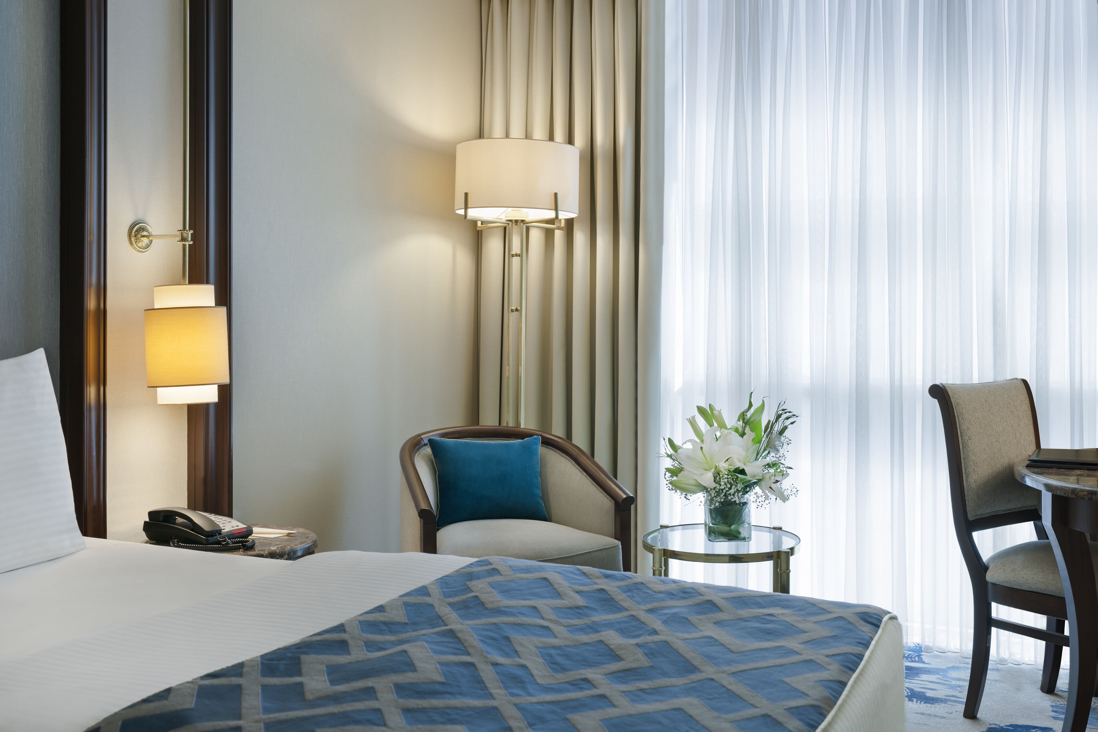 A hotel room with a bed, two chairs, a small table with a vase of flowers, a lamp, and a telephone on the nightstand.