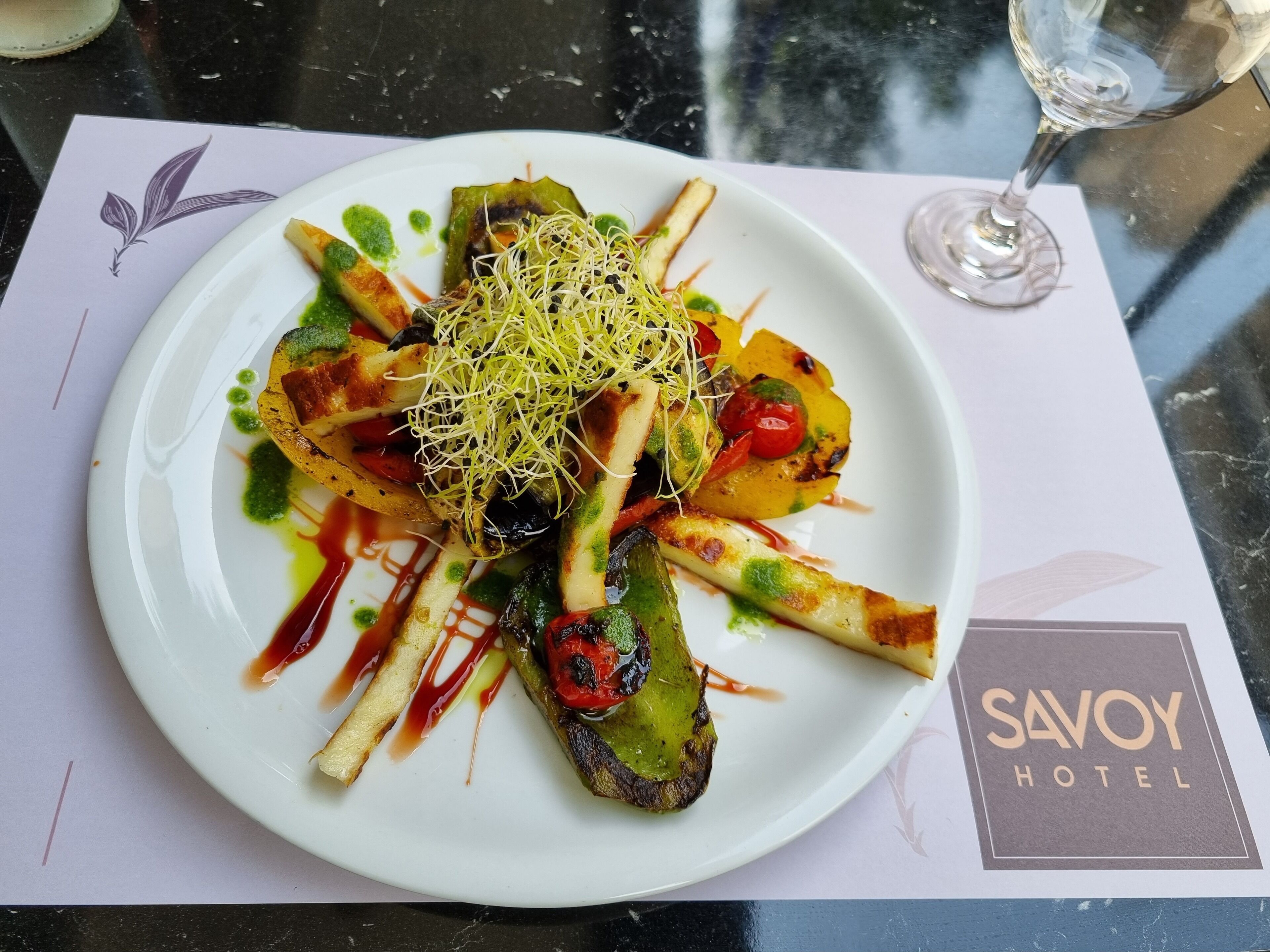 Prato de comida com um copo de vinho sobre uma mesa com jogo de toalha do Savoy Hotel.