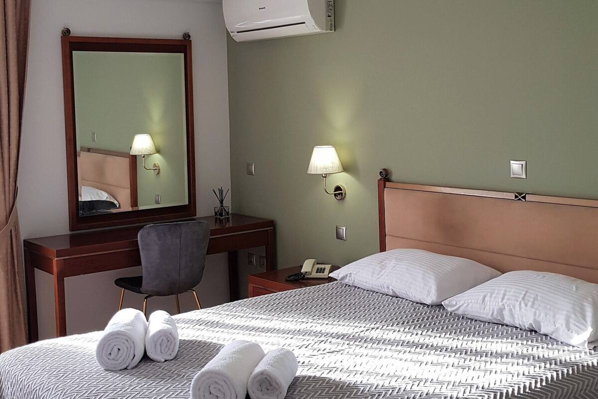 Habitación de hotel con cama, escritorio, silla, espejo, aire acondicionado de pared y dos lámparas de pared.