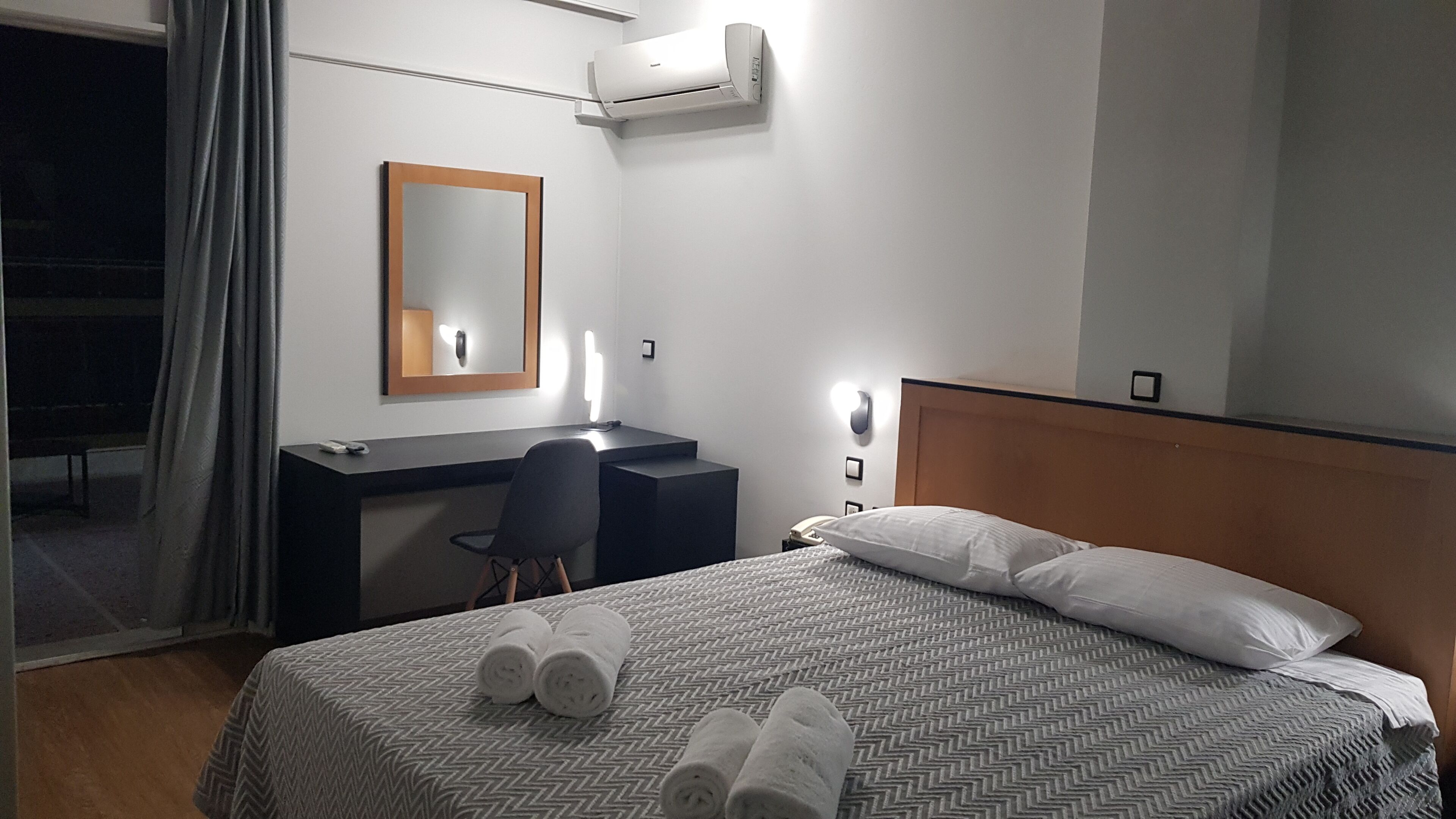 A hotel room with a bed, a desk, a chair, a mirror, and an air conditioning unit.