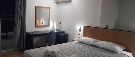 A hotel room with a bed, a desk, a chair, a mirror, and an air conditioning unit.