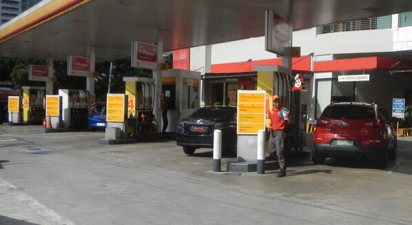 Une station-service avec plusieurs pompes à essence, une personne debout près d’un véhicule rouge et un véhicule bleu garé à l’arrière-plan.
