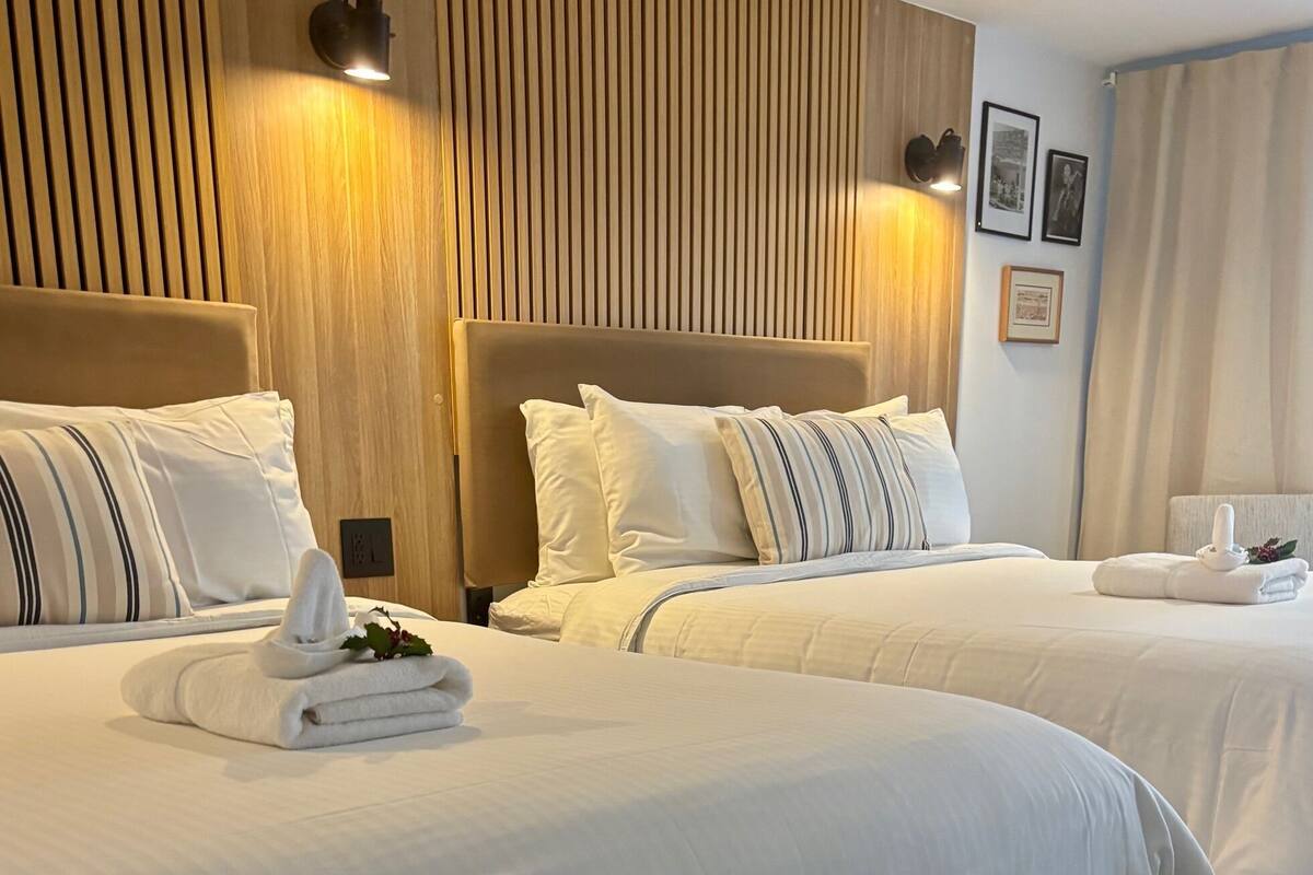 Habitación de hotel con dos camas, cada una con ropa blanca y almohadas a rayas. Hay apliques de pared y cuadros enmarcados.