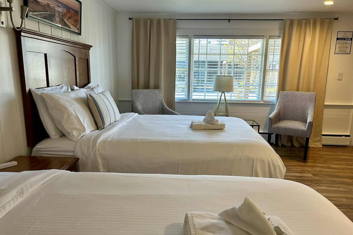 Habitación de hotel con dos camas, cabecero de madera, ventana con persianas, una silla y una lámpara.
