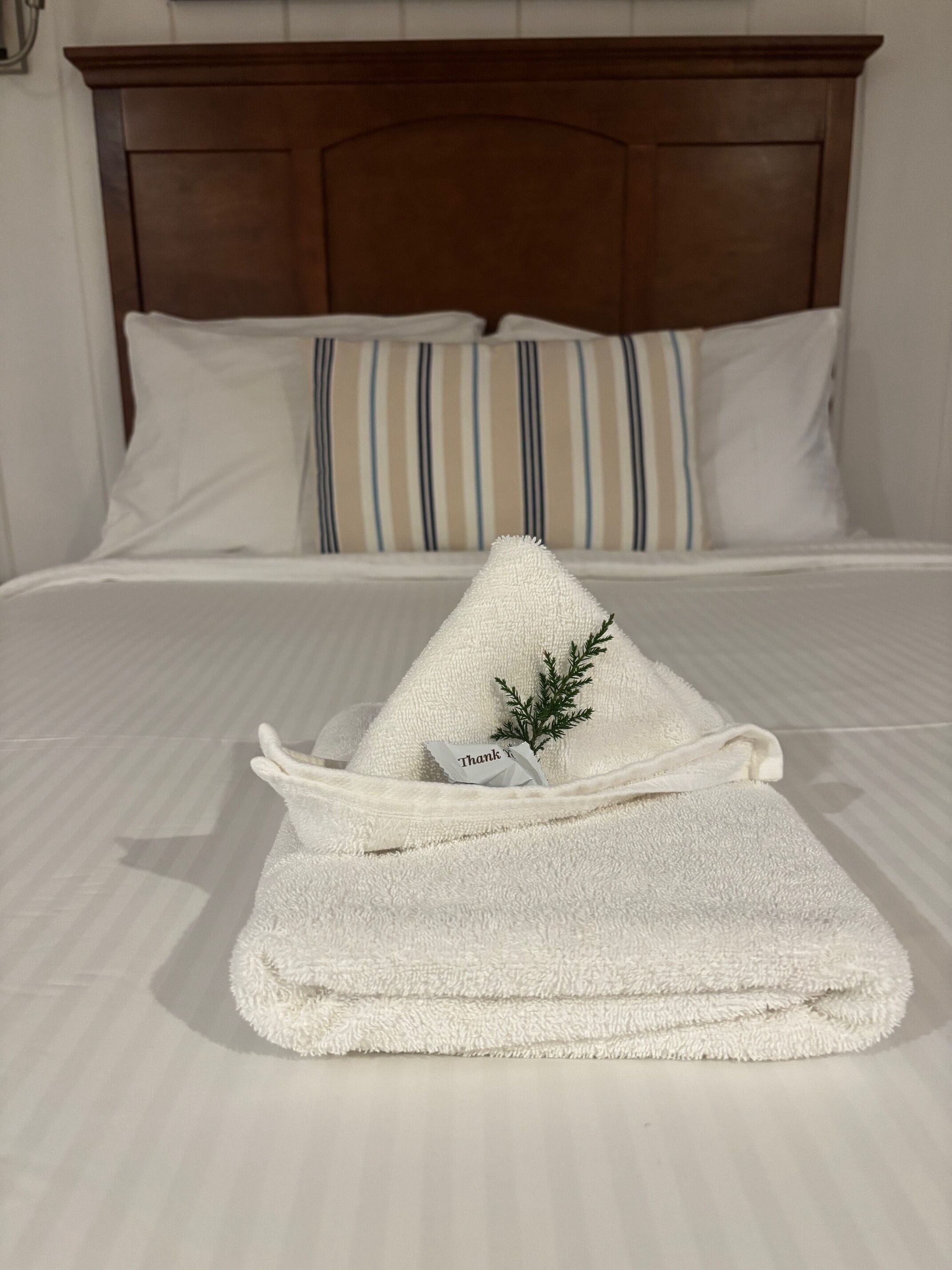 A neatly made bed with white bedding, striped pillows, and a folded towel with a 'Thank You' note and a sprig of greenery.