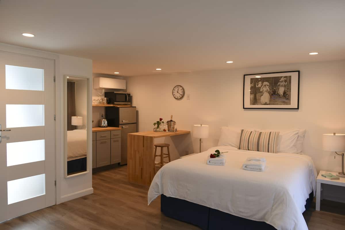 Habitación de hotel con cama, mesita de noche, escritorio, kitchenette y un reloj en la pared.