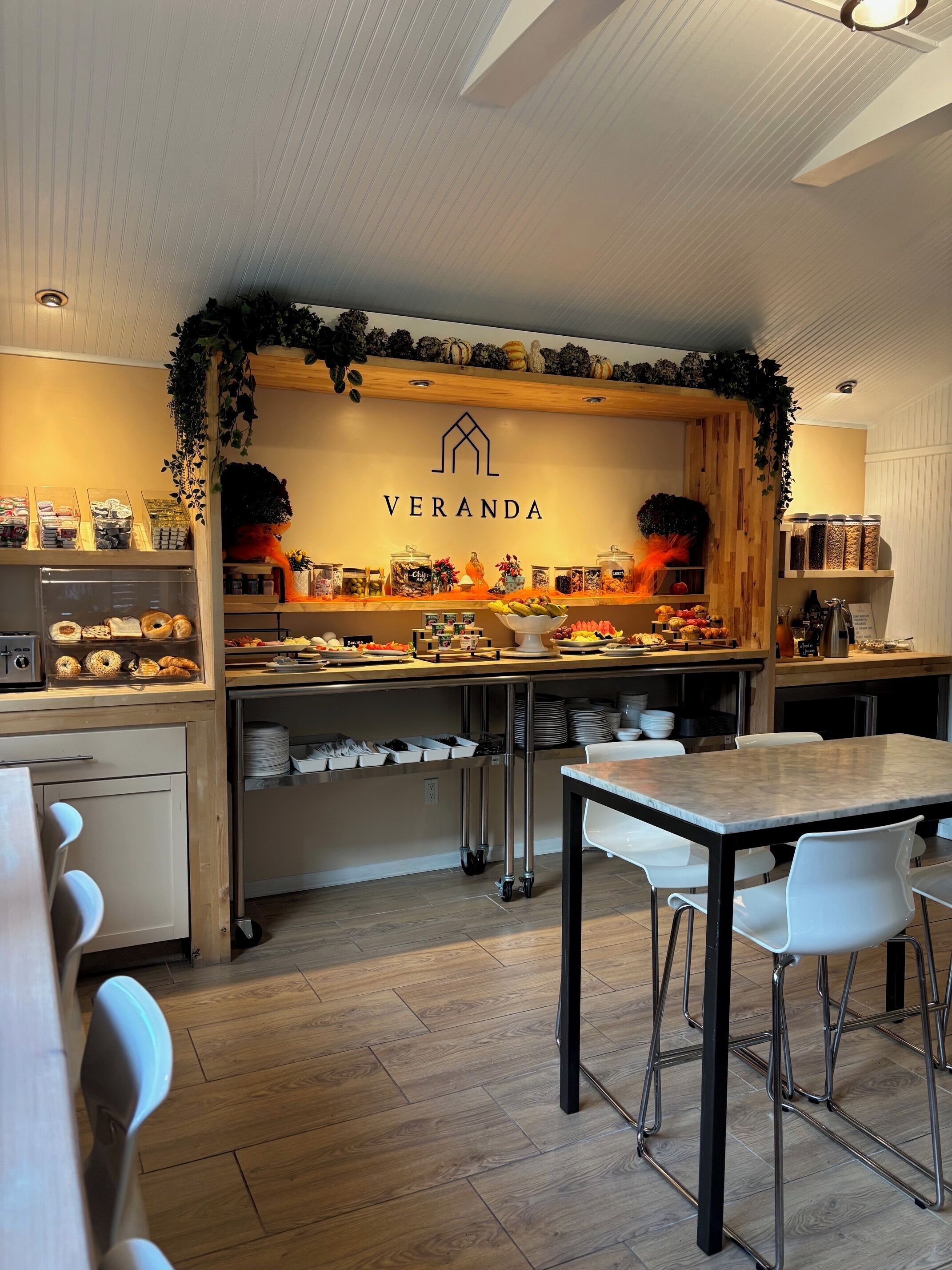 Een café-interieur met een houten toonbank waarop diverse etenswaren staan, een bord met de tekst 'VERANDA' en een zithoek met witte stoelen en een tafel.