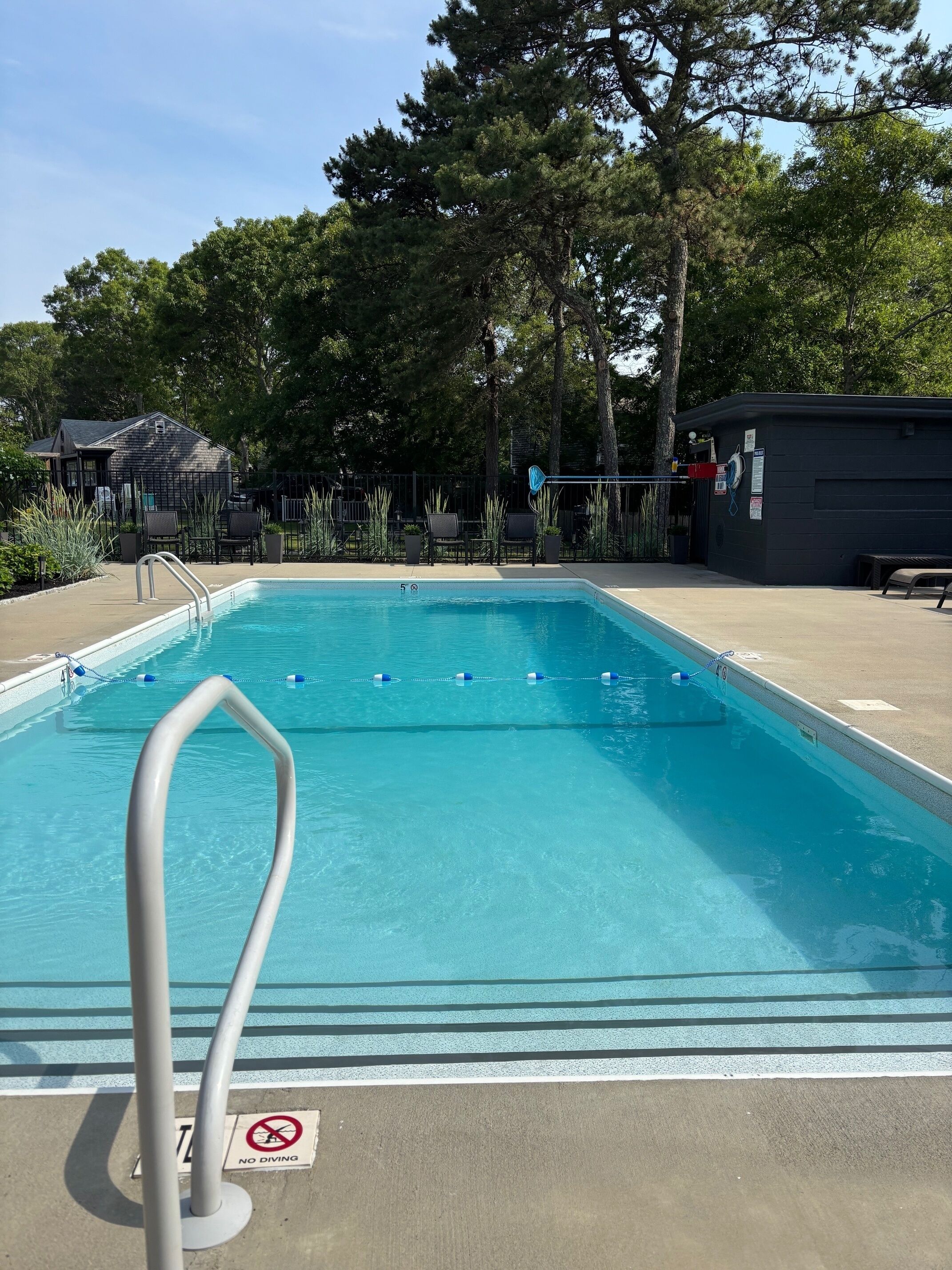 Une piscine aux eaux cristallines, agrémentée d’un panneau indiquant qu’il est interdit de plonger, entourée d’arbres et bordée d’une zone délimitée par une clôture, où se trouvent des sièges pour profiter de l’extérieur.