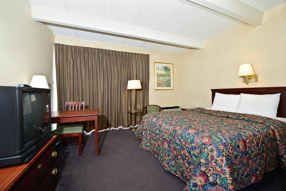 A hotel room with a bed, a desk with a chair, a television, a lamp, and a painting on the wall.