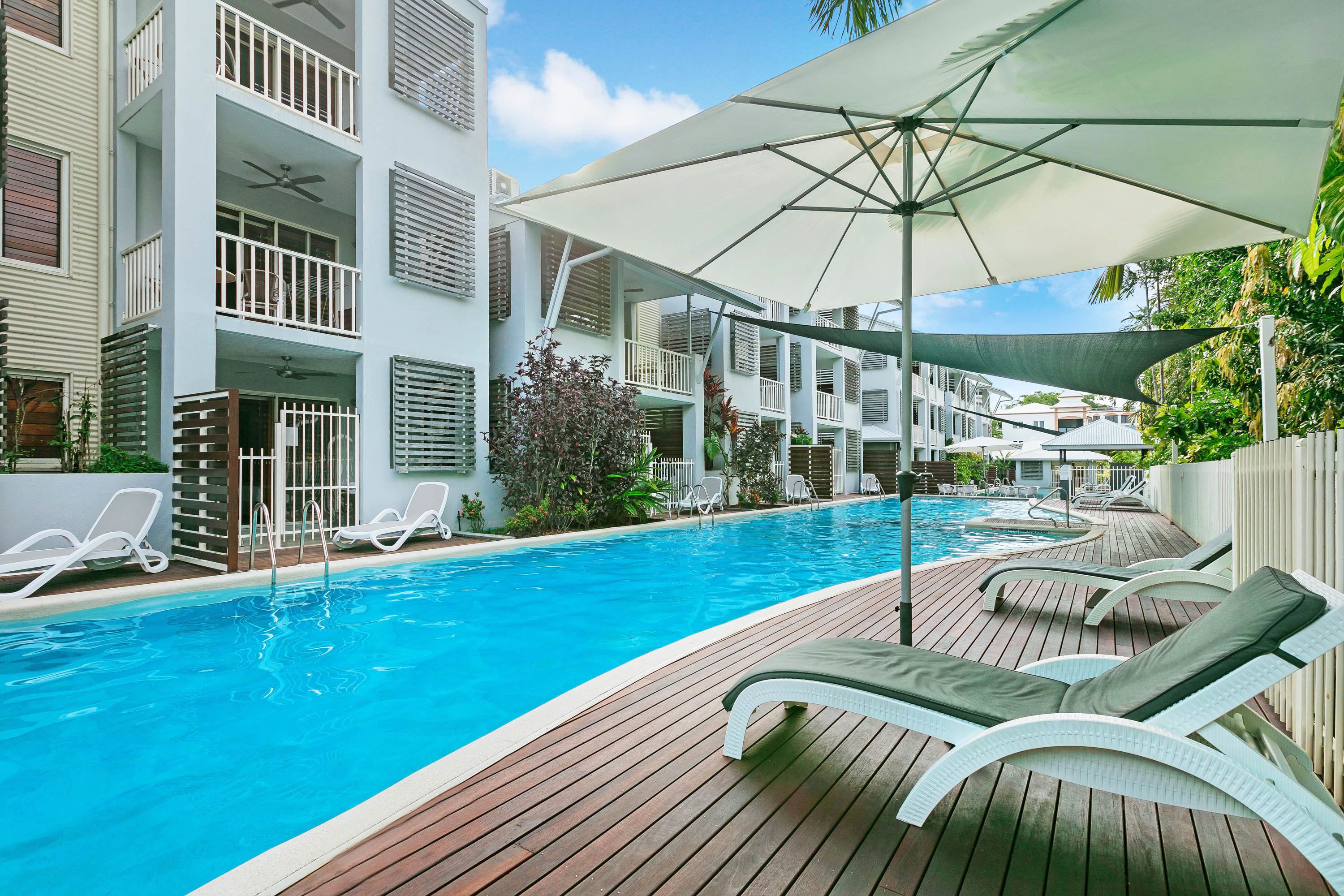 A swimming pool with lounge chairs and umbrellas, surrounded by a wooden deck and a white building with balconies.