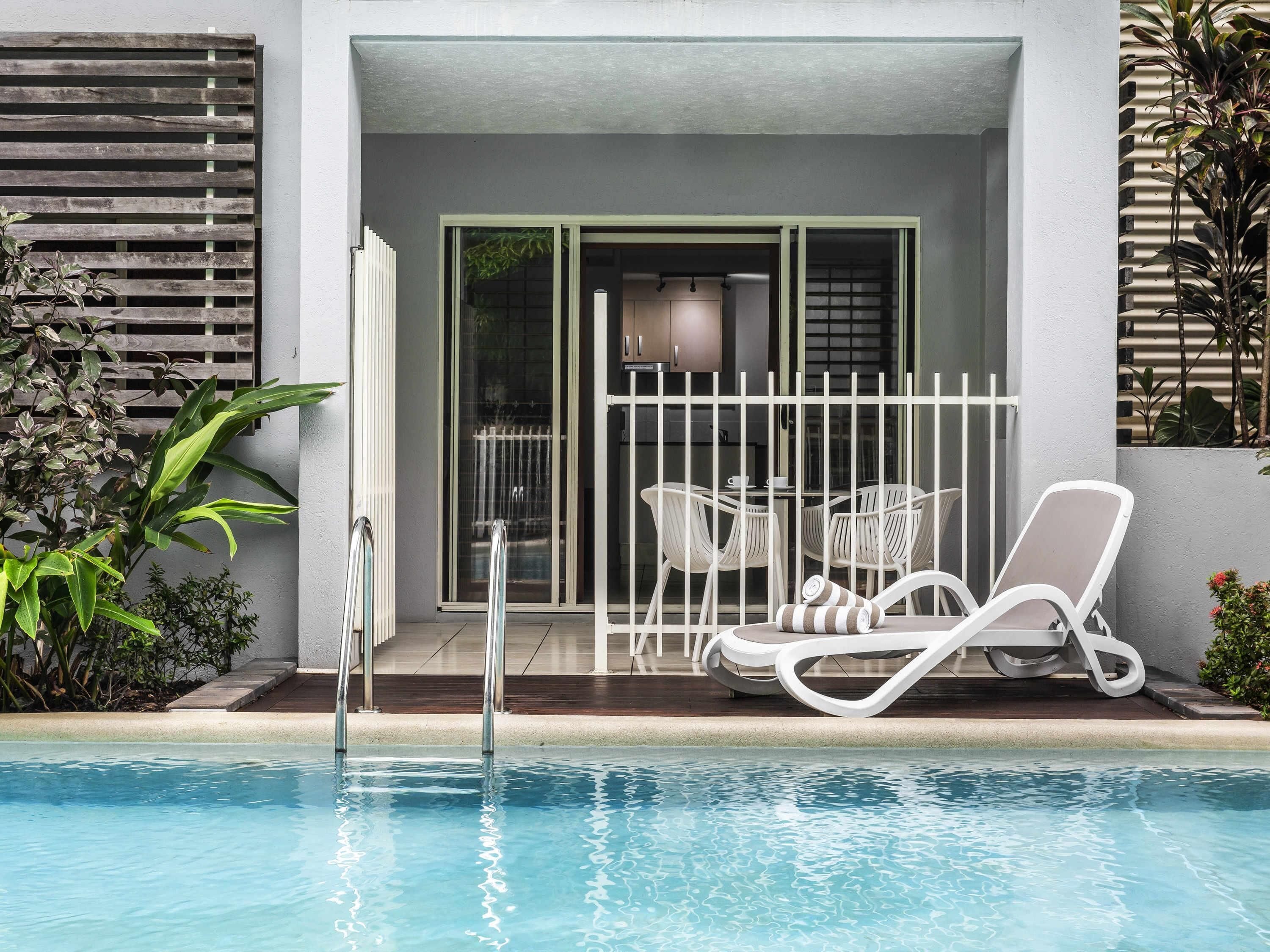 A poolside area with a glass door leading to a room, white lounge chairs, and a small table.