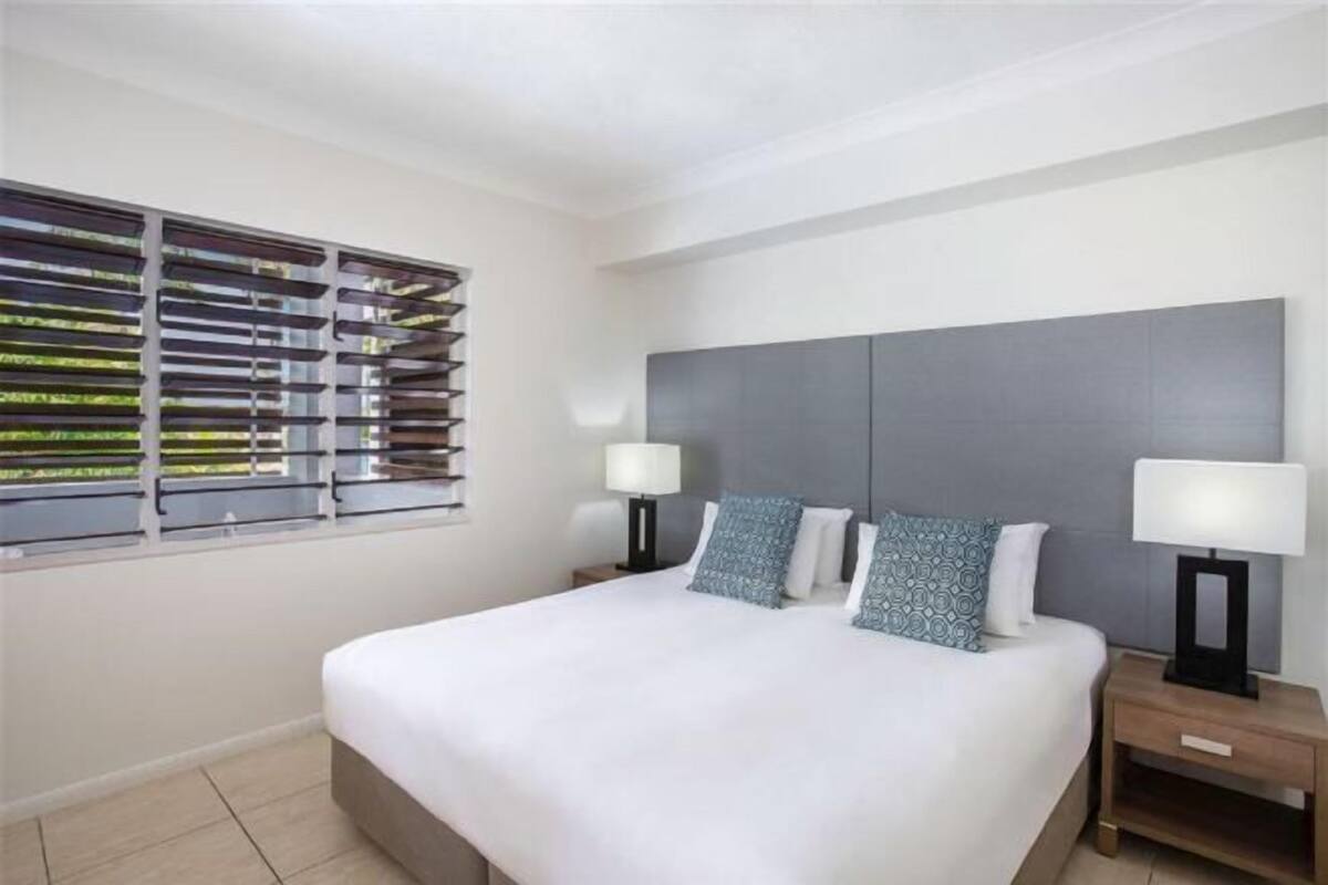 A hotel room with a large bed, two bedside tables, a window with blinds, and a grey headboard.