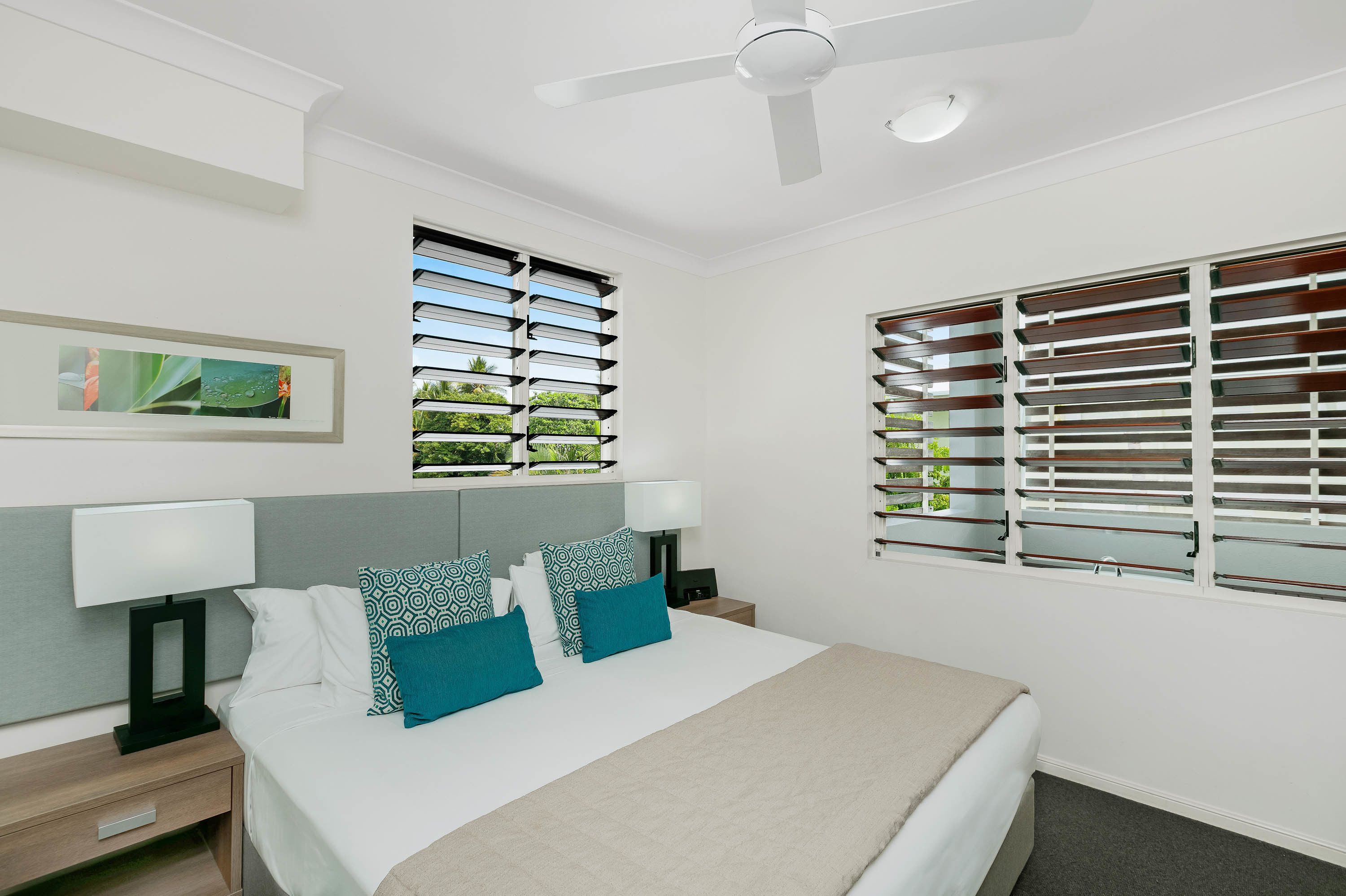 Quarto de hotel moderno com uma cama grande, duas mesas de cabeceira com luminárias, um ventilador de teto e duas janelas com persianas.