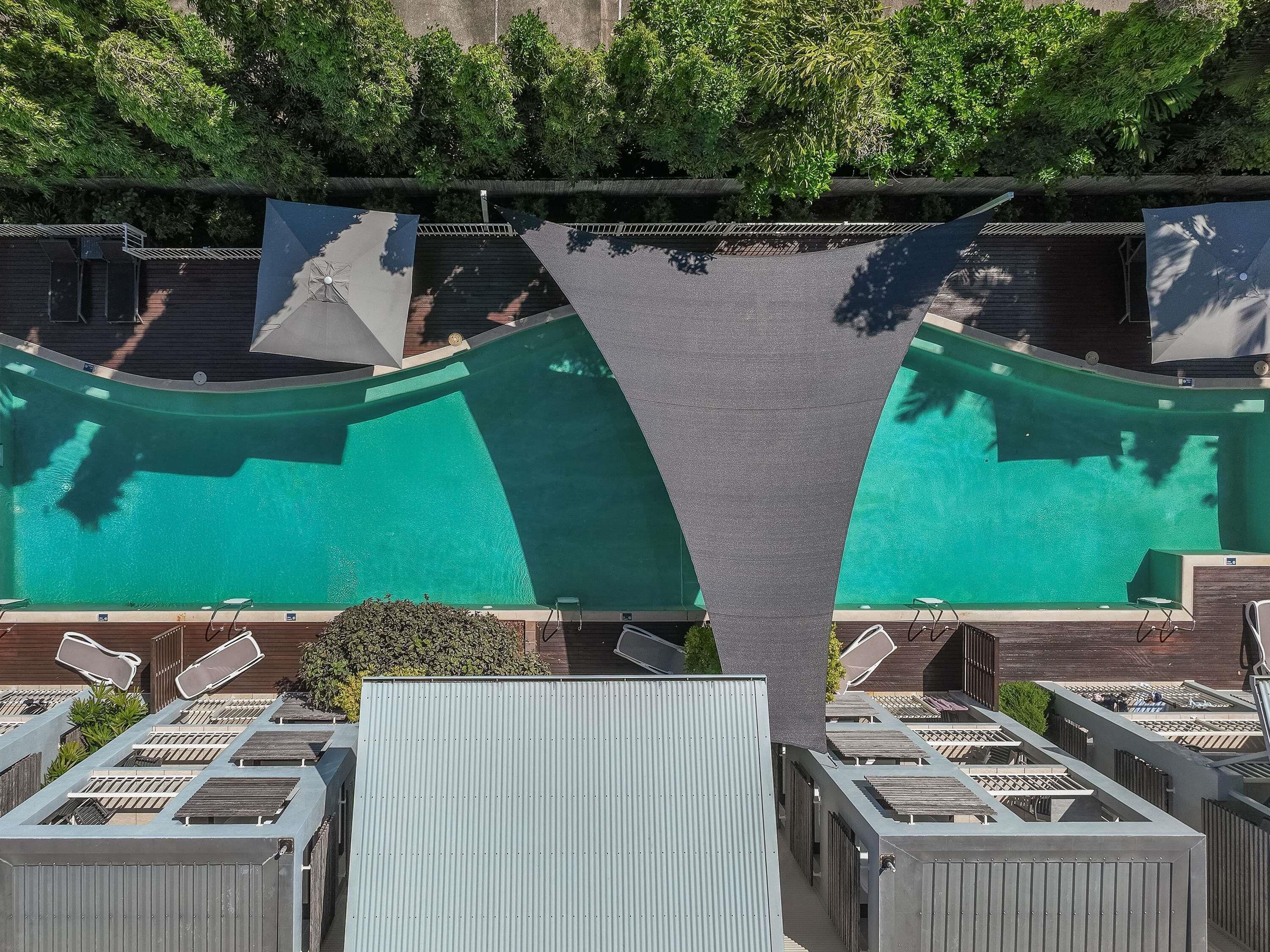 A swimming pool with a wooden deck, surrounded by metal railings and shaded by large umbrellas.
