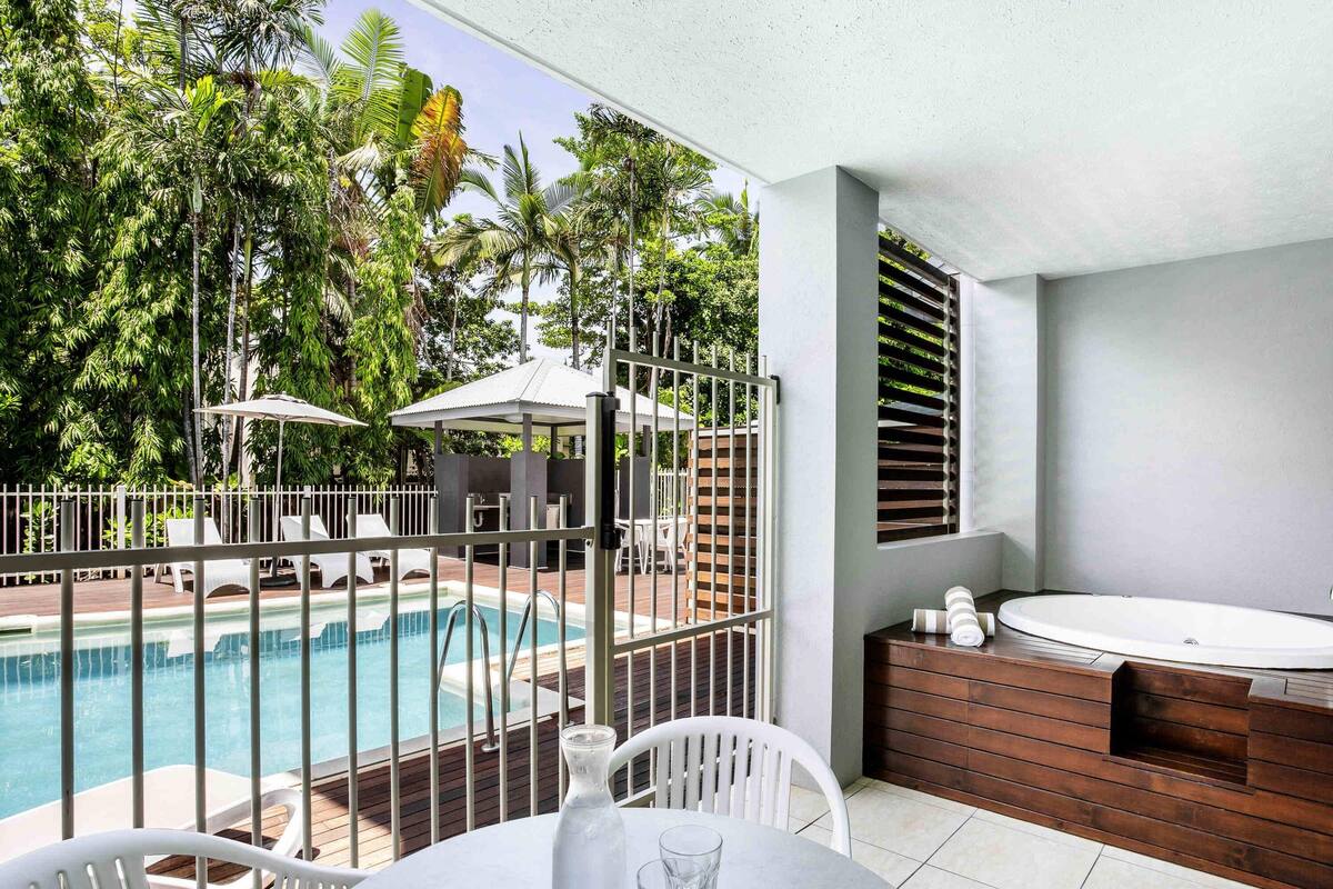 A poolside area with a view of the pool, a table with chairs, a wooden bench, and a window with blinds.