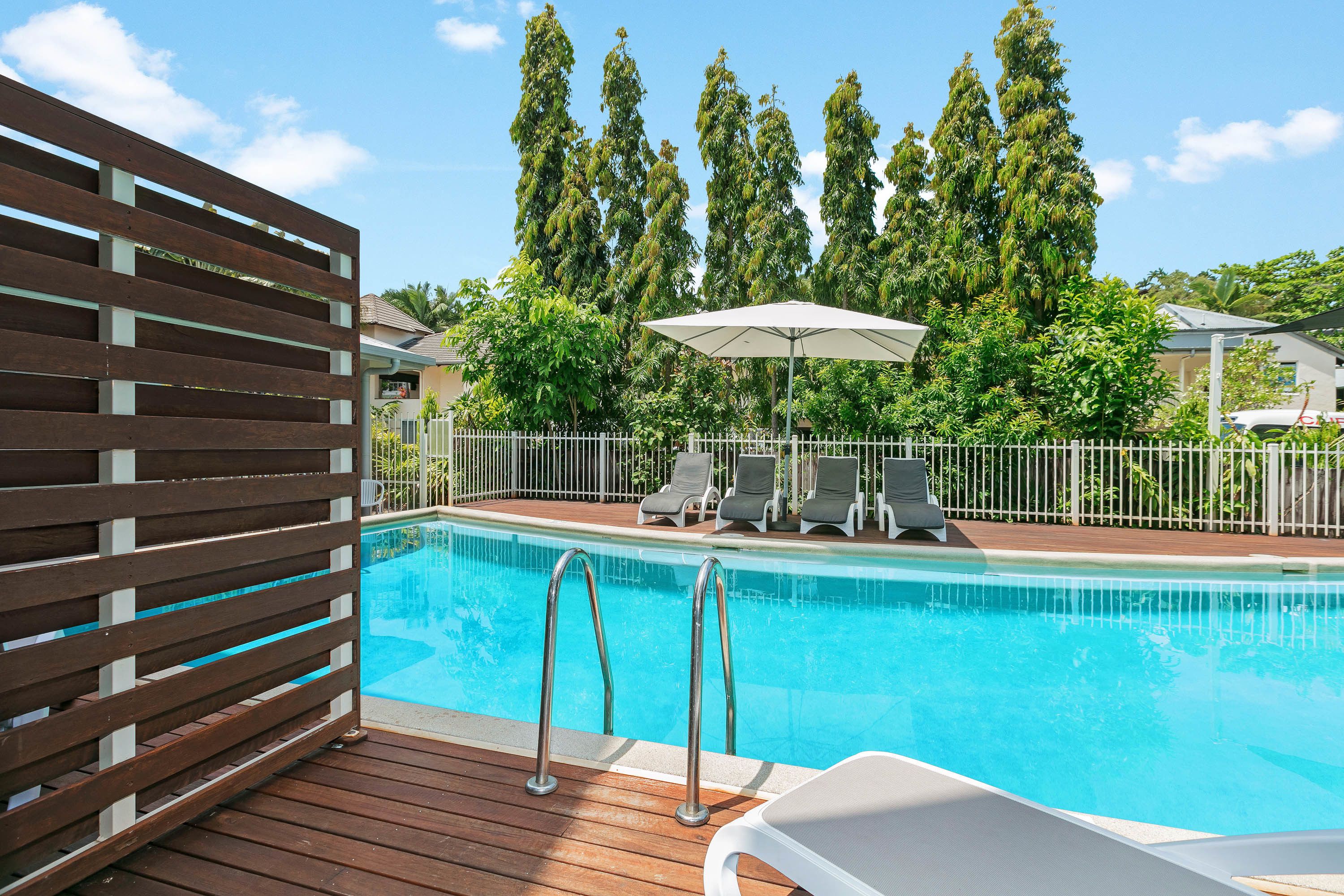 Piscina con sillas de descanso y sombrillas, rodeada por una terraza de madera y una valla blanca.