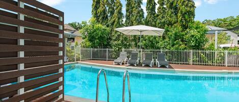 Piscina con sillas de descanso y sombrillas, rodeada por una terraza de madera y una valla blanca.
