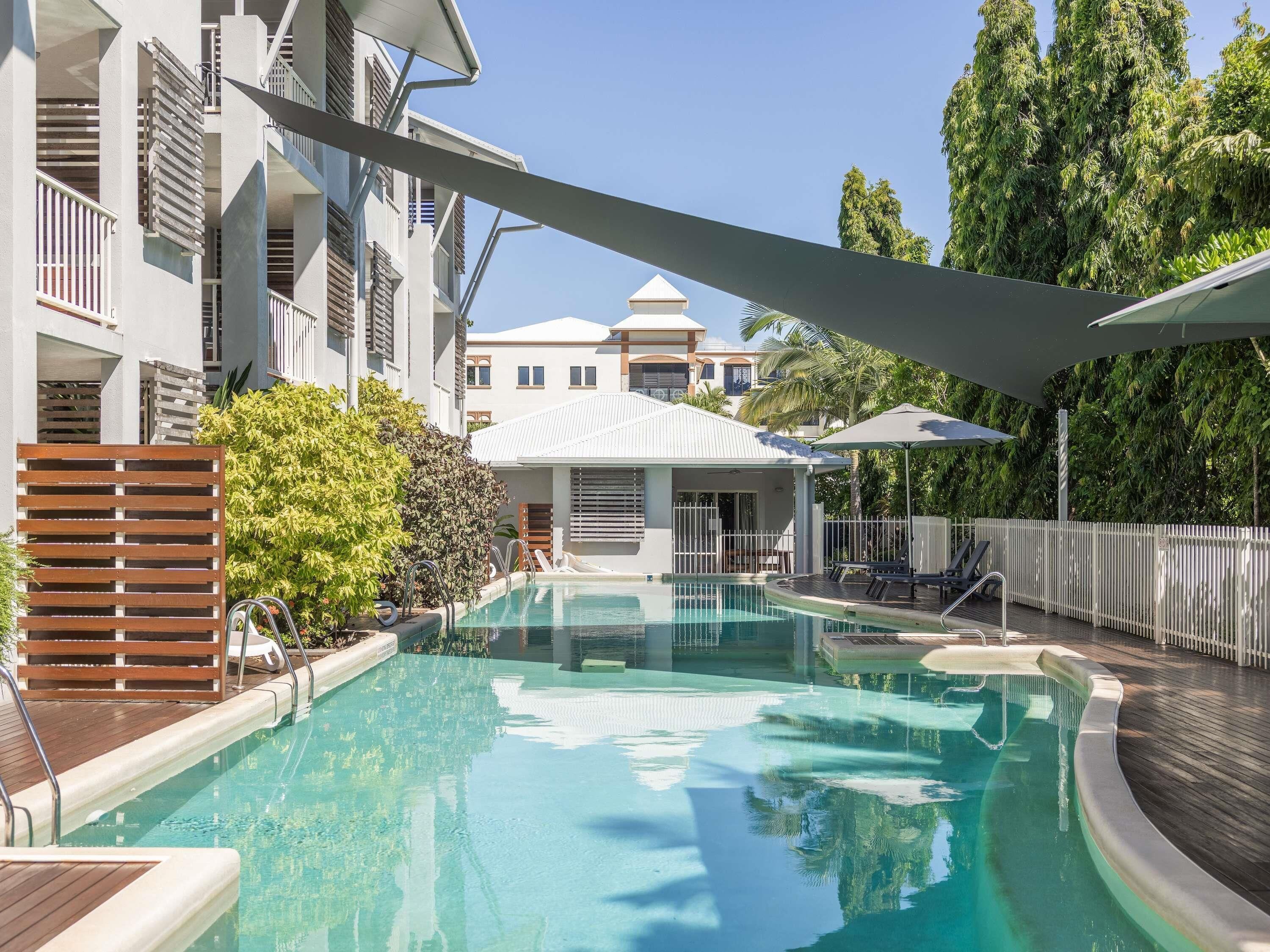A swimming pool with a wooden deck, surrounded by buildings with balconies and a covered area.