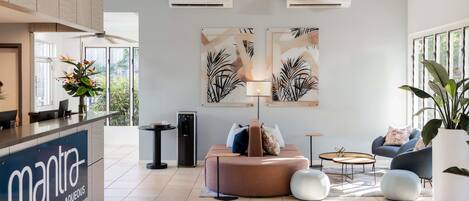 A modern hotel lobby with a reception desk, seating area, ceiling fan, air conditioning units, and artwork on the walls.