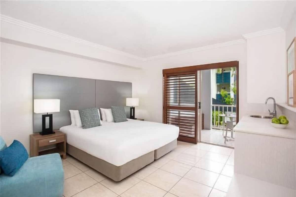 A bedroom with a large bed, bedside tables, a nightstand, a sofa, a balcony with a view, and a kitchenette.