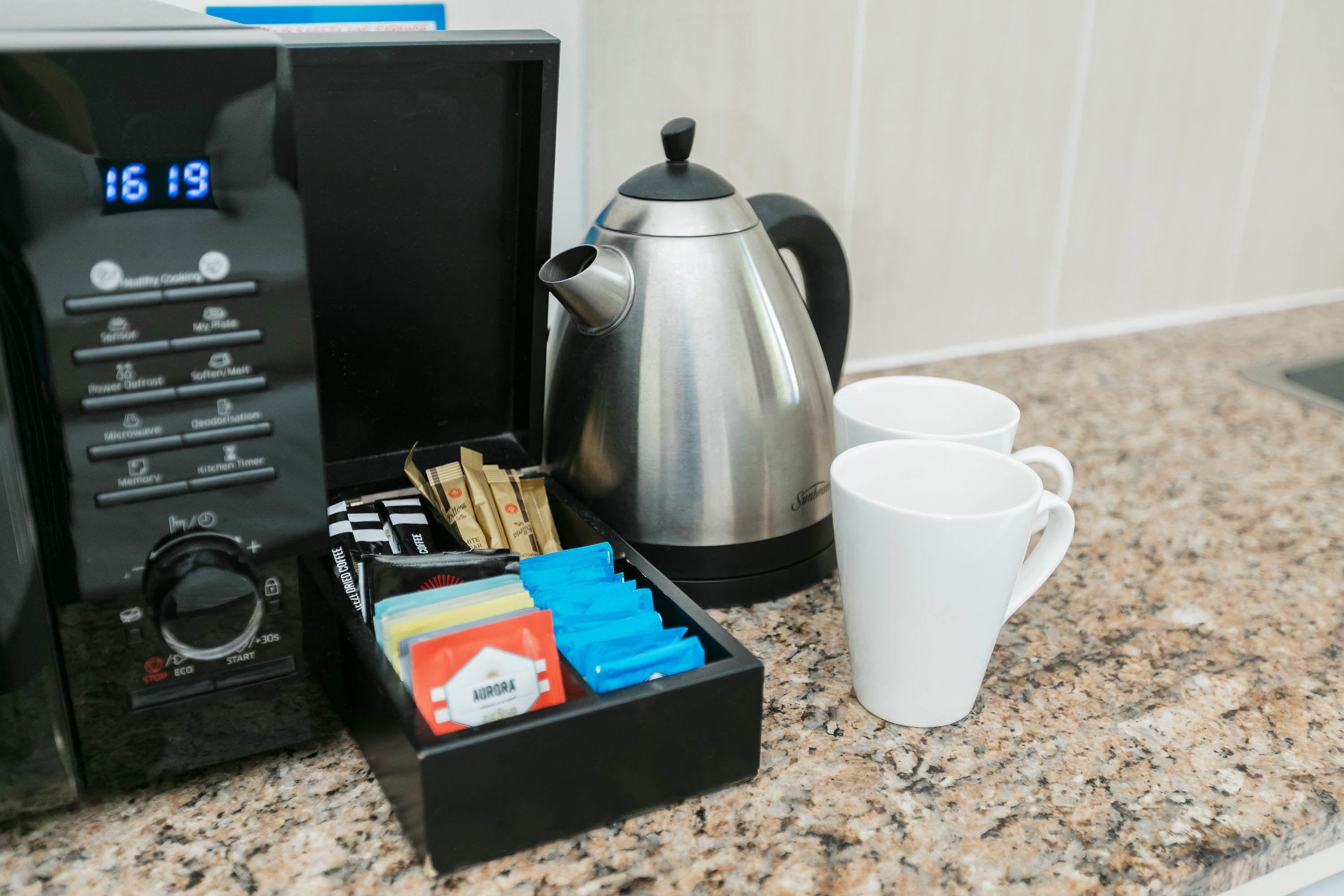 Un microondas con pantalla digital, una tetera eléctrica de acero inoxidable, una taza blanca y un cajón con varios objetos, incluyendo una caja roja con la etiqueta "AURORA".