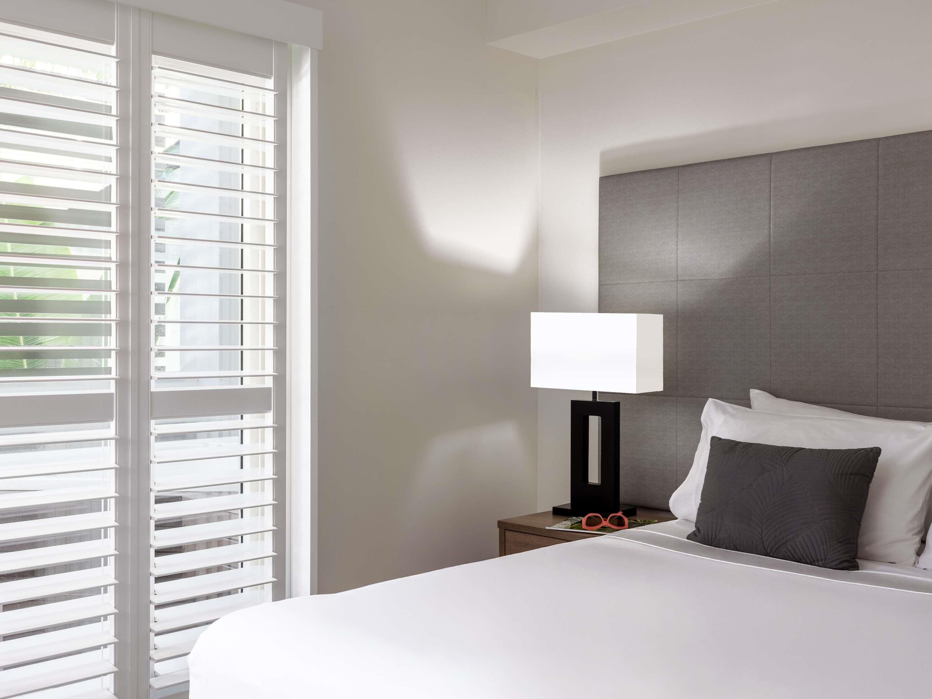 A hotel room with a bed, a nightstand, a lamp, and a window with blinds.