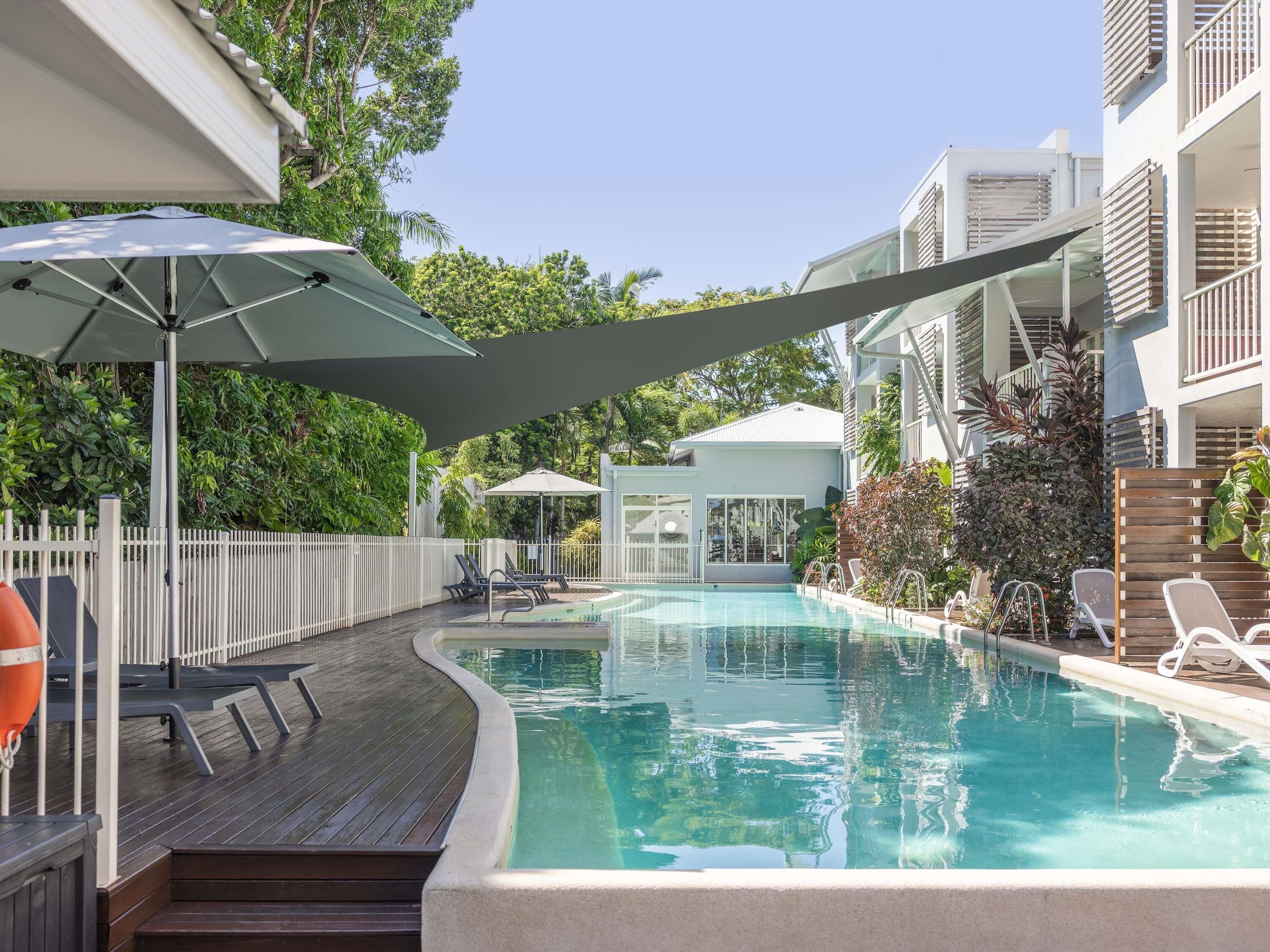 A swimming pool with lounge chairs and umbrellas, surrounded by a wooden deck and a white fence.