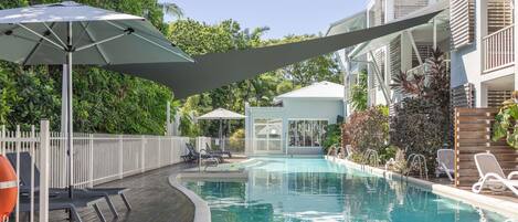 A swimming pool with lounge chairs and umbrellas, surrounded by a wooden deck and a white fence.