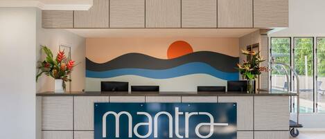 Un comptoir d’accueil avec une enseigne indiquant « mantra AQUEOUS », une fresque murale représentant un coucher de soleil et une vue sur l’espace piscine.
