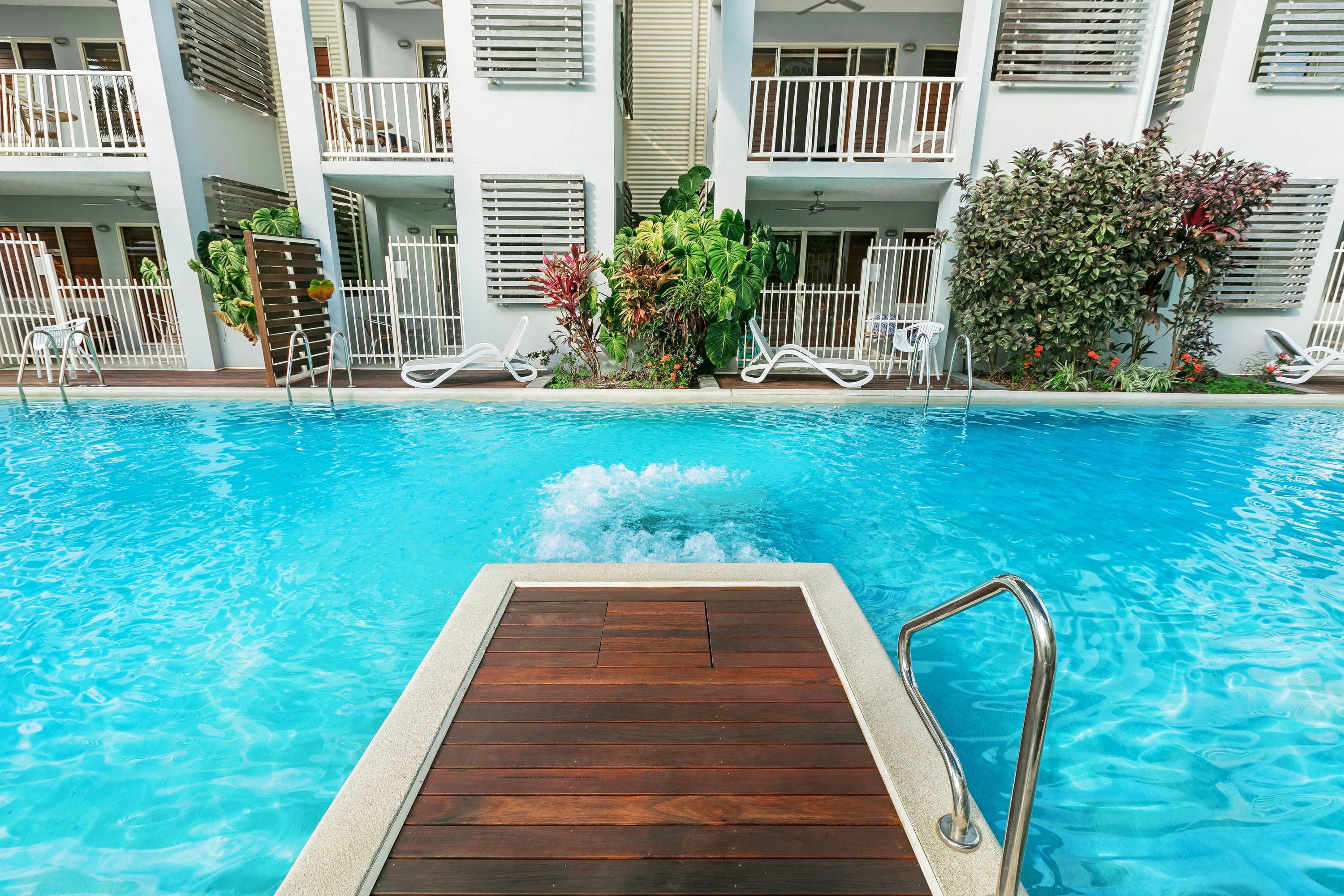 A swimming pool with a wooden deck and metal railings, surrounded by white balconies with railings and plants.