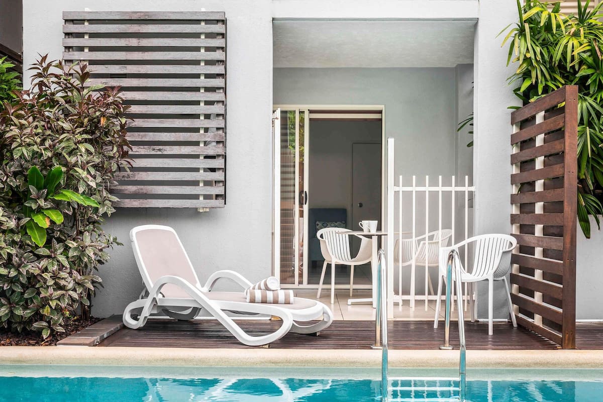A poolside area with lounge chairs and a table, a wooden fence, and a door leading to an interior space.