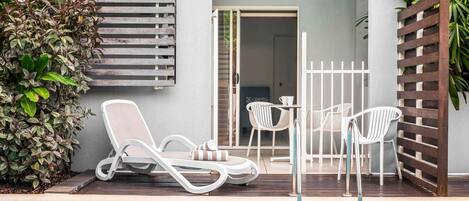 Zona de piscina con sillas reclinables y una mesa, una valla de madera y una puerta que da a un espacio interior.