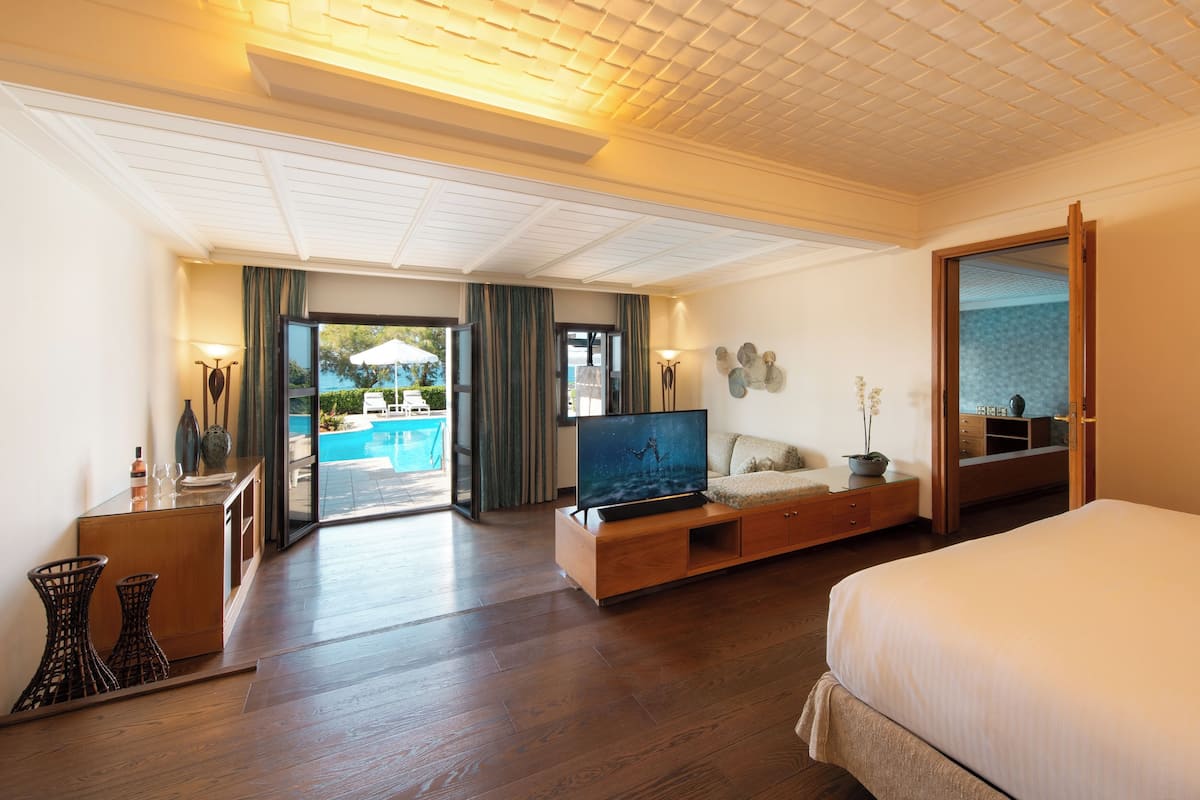 Une chambre d’hôtel avec un grand lit, un téléviseur posé sur un meuble en bois, une armoire en bois, une petite table avec une lampe, et une vue sur une piscine à travers une porte vitrée.