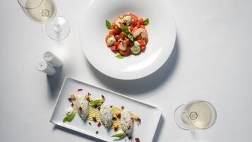 A white plate with a salad and a rectangular dish with food items, accompanied by two glasses of white wine on a white table.