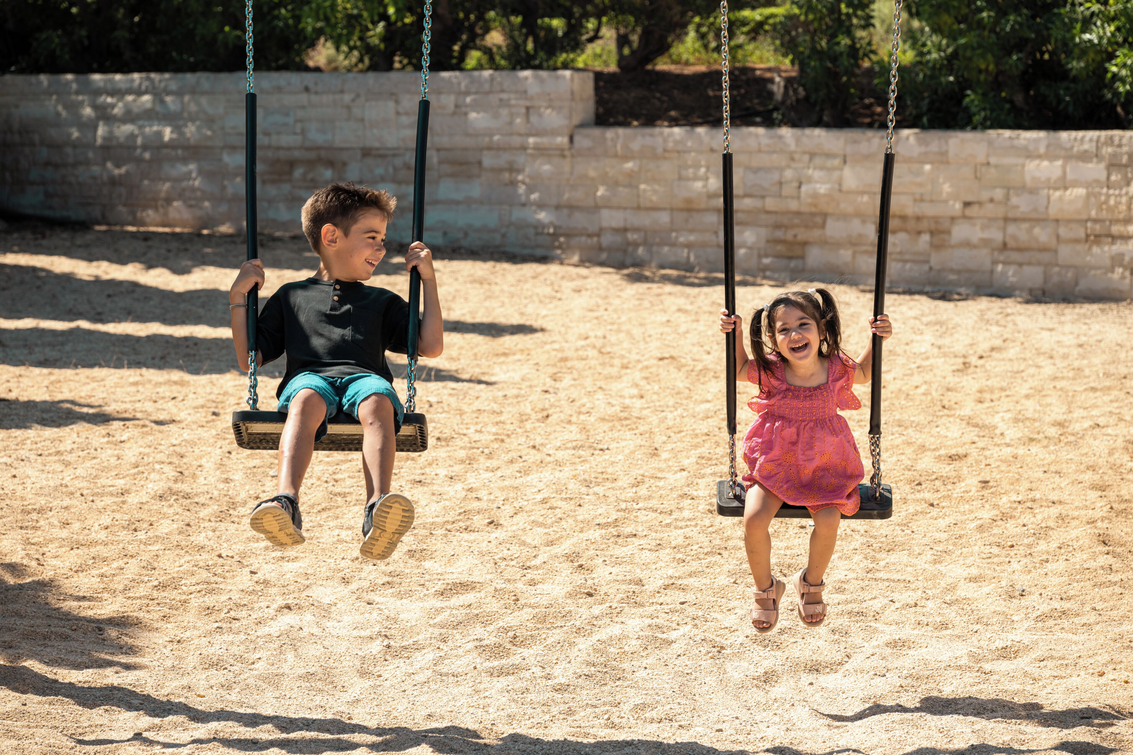 Two children on swings in a sandy area with a stone wall in the background.