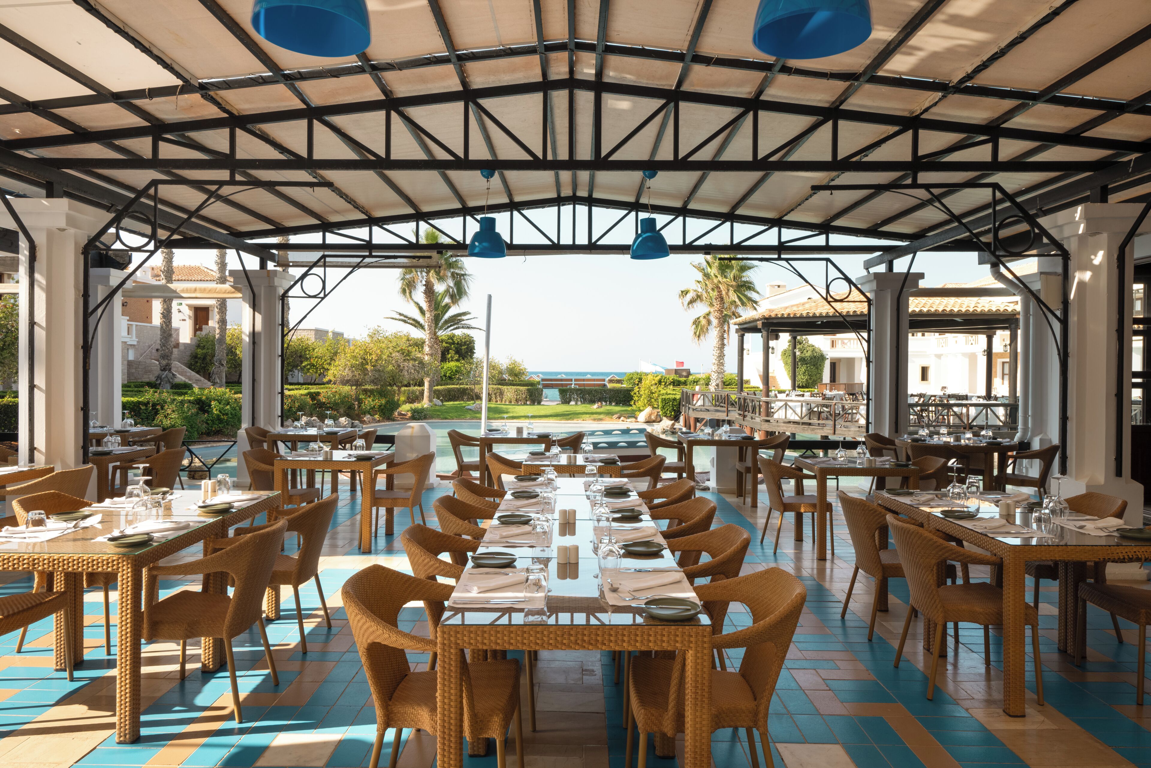 An outdoor dining area with tables and chairs, a covered roof, and a view of the sea.