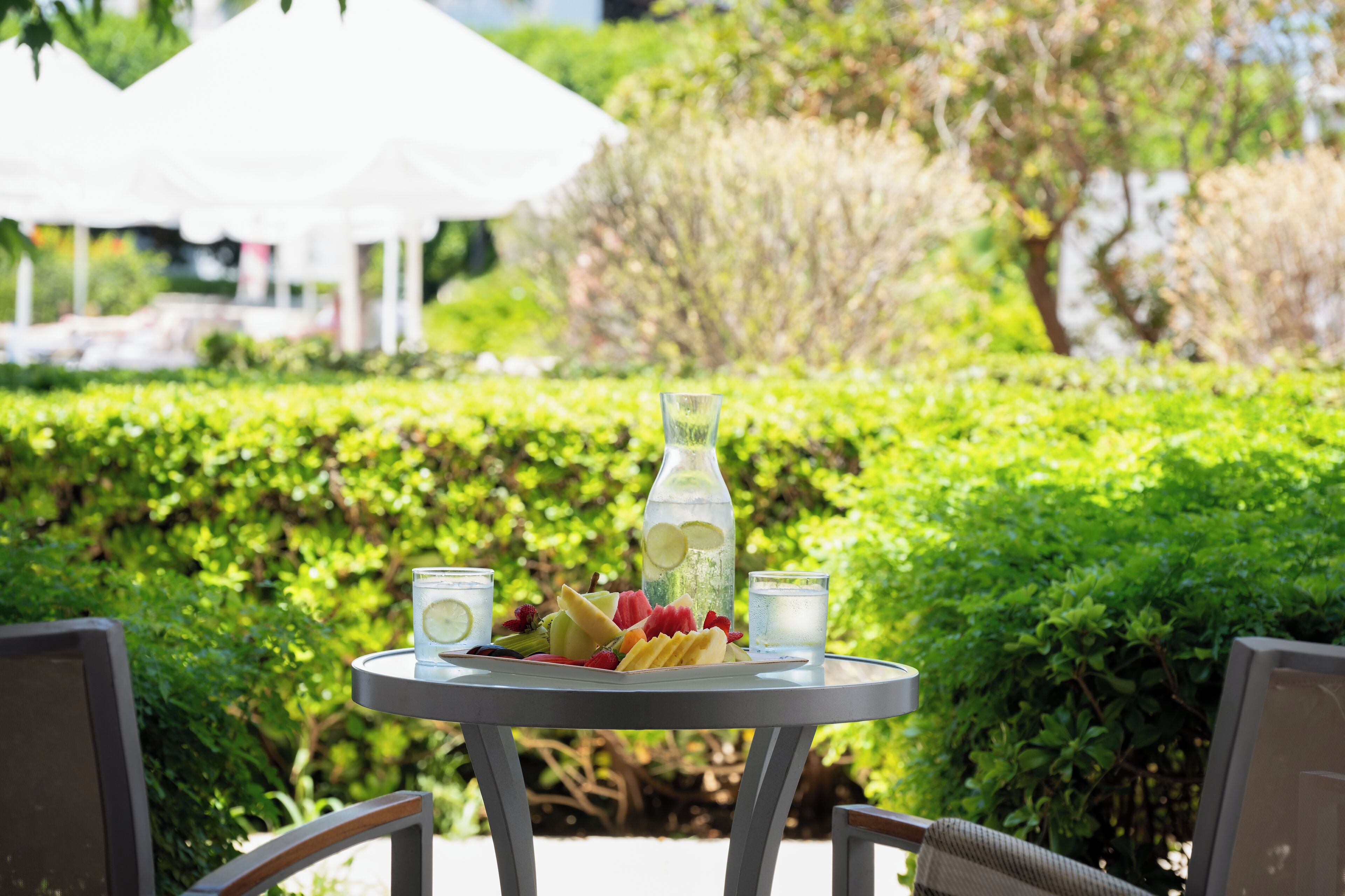A round table with a bottle, glasses, and a fruit platter in an outdoor setting with greenery and white umbrellas in the background.