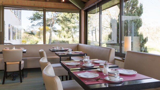 Un restaurant avec des tables dressées pour le service, de grandes fenêtres et une vue sur l’extérieur.
