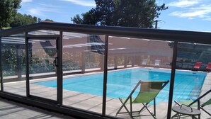 Une piscine extérieure couverte, dotée d’une structure en verre, d’un revêtement de sol carrelé et de quelques chaises longues.