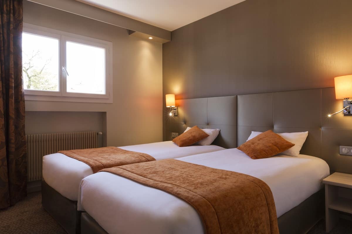 Une chambre d’hôtel avec deux lits, une fenêtre, un radiateur et une table de chevet avec une lampe.