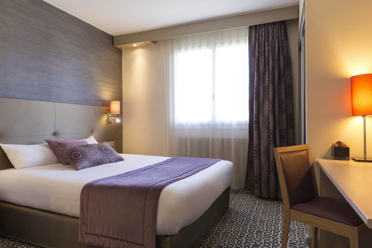 Une chambre d’hôtel avec un lit, un bureau, une chaise, une lampe et une fenêtre avec des rideaux.
