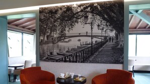 Une pièce avec deux fauteuils orange, une table ronde sur laquelle se trouve un plateau avec des tasses et des soucoupes, et une grande fresque murale en noir et blanc représentant un paysage au bord de la rivière.